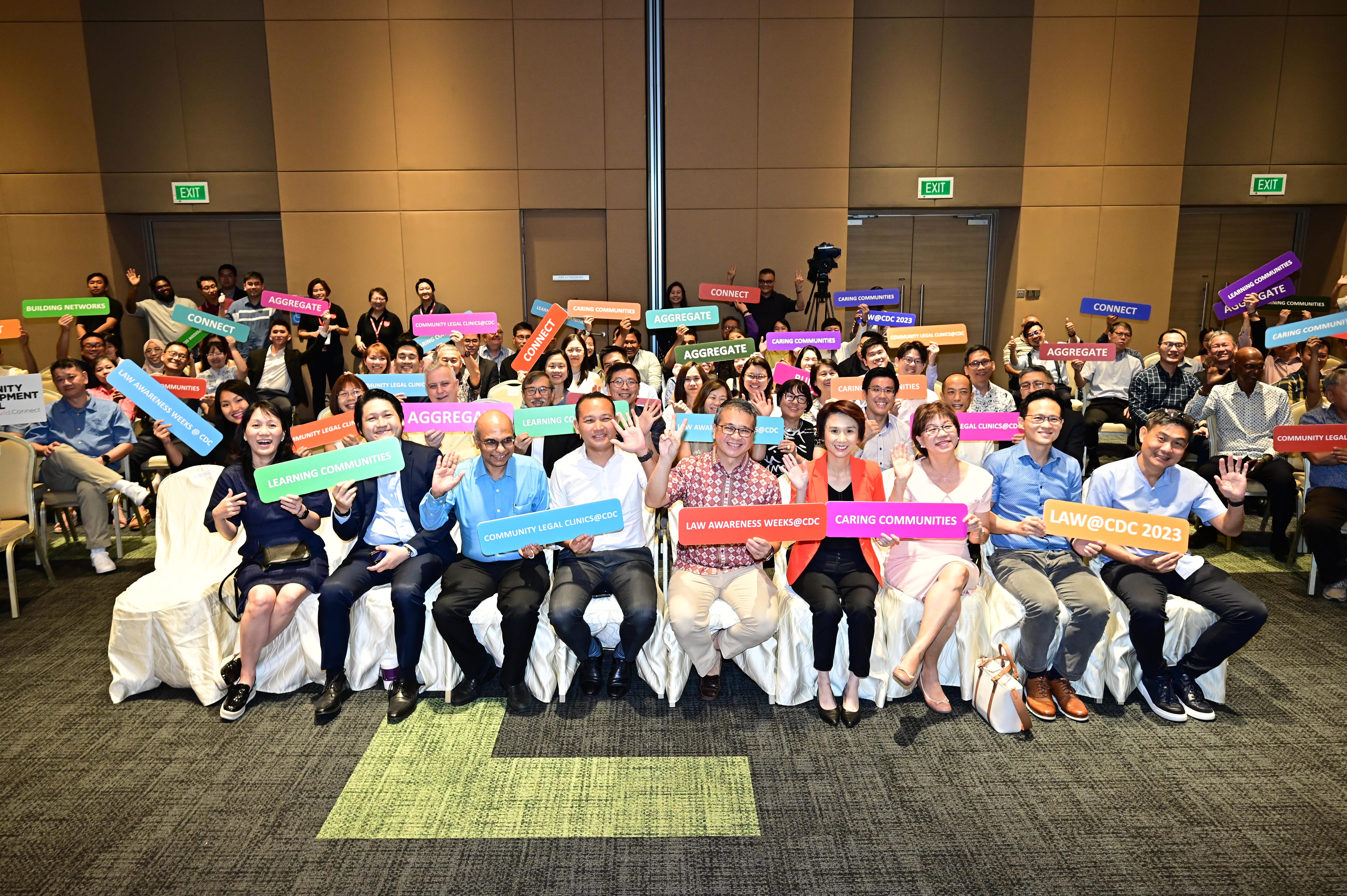 People seated in a large room holding colorful signs with various phrases like "CONNECT," "AGGREGATE," and "COMMUNITY LEGAL CLINICS."